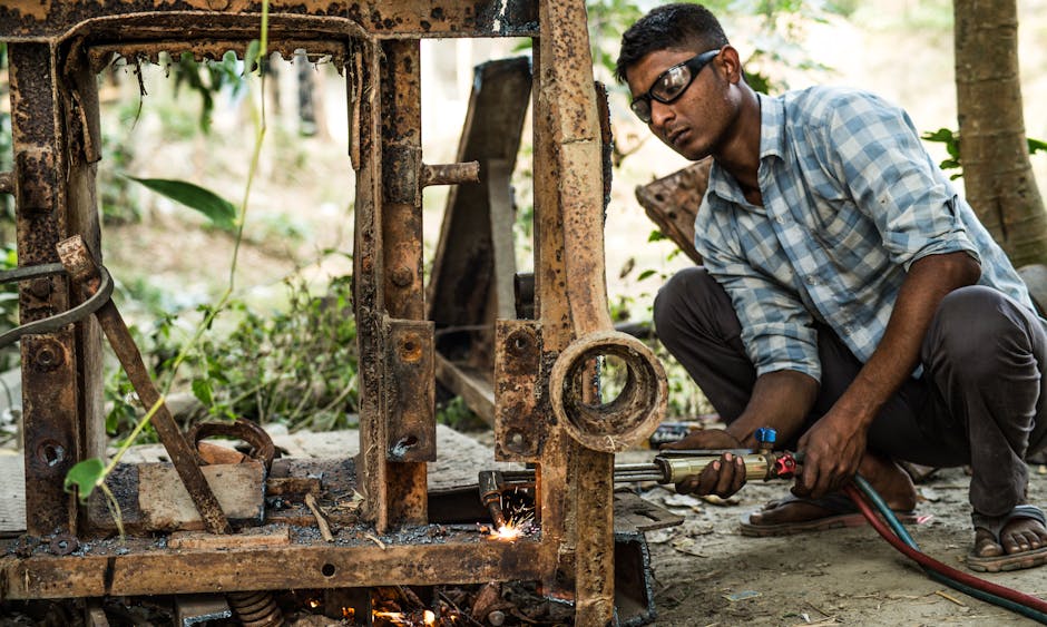 An adult male welder focused on repairing a rusty metal machine outdoors using a welding torch.